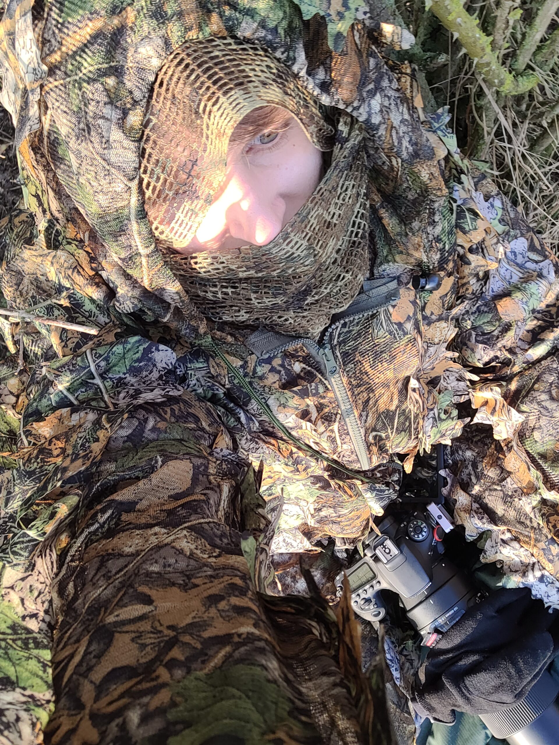 Person liegt in Tarnkleidung am Boden mit Kamera für Naturfotografie.