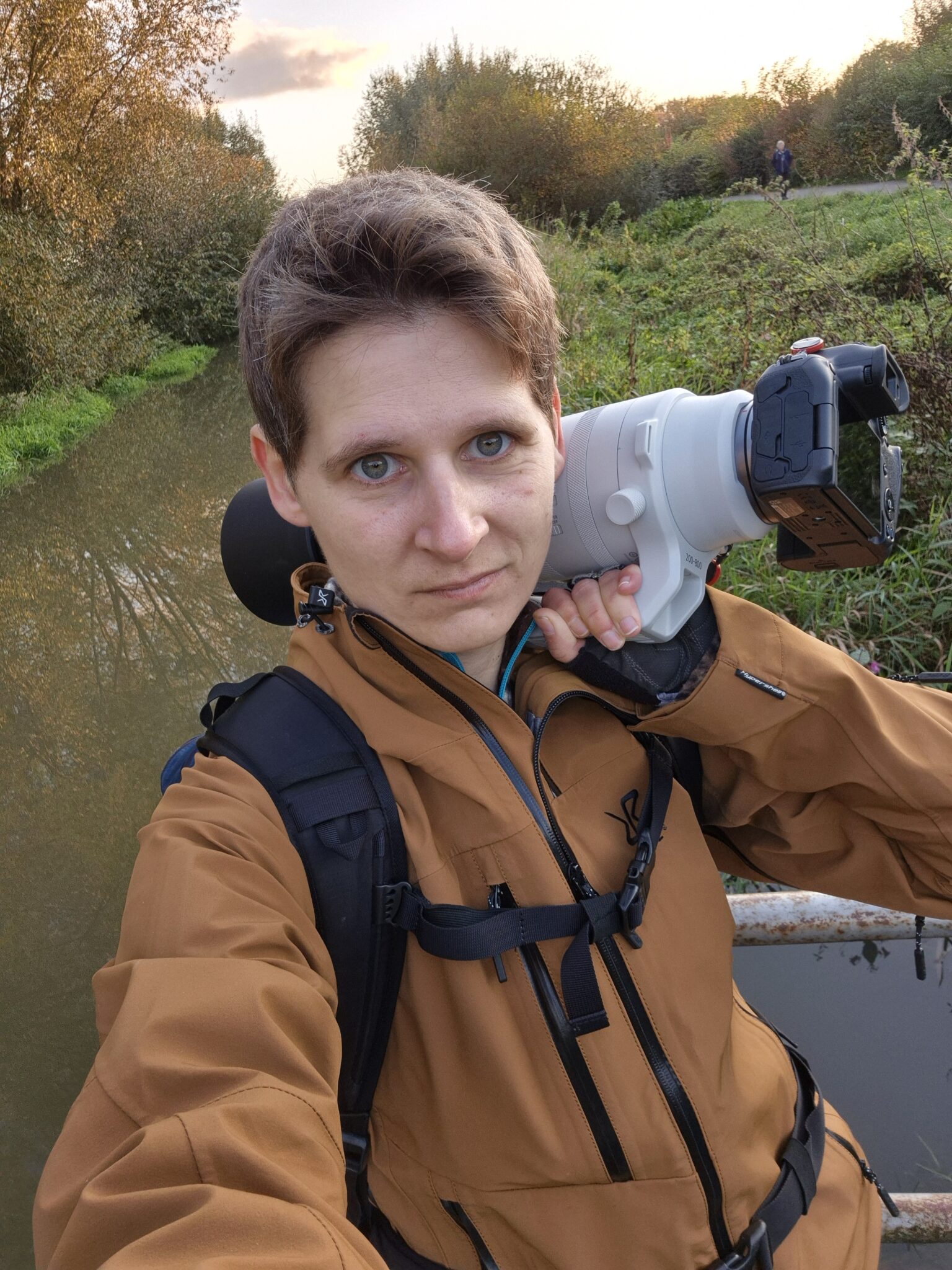 Person mit großem Teleobjektiv steht an einem Gewässer und macht ein Selfie.